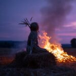 Wächterfigur aus Stroh vor lodernden Flammen unter violettem Abendhimmel – Ein Sinnbild für Pflanzengeister und Naturmagie.